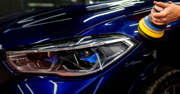 A close-up of a dark blue car being polished with a yellow pad, highlighting its sleek headlight and reflective surface.