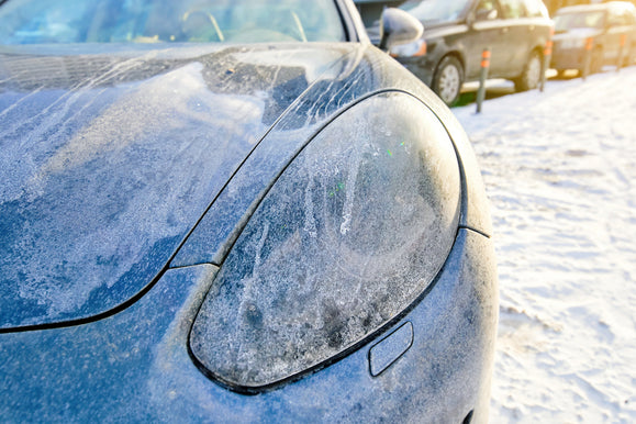 A close-up of a frosty car headlight and hood, covered in road salt and ice, parked in line at a car wash.