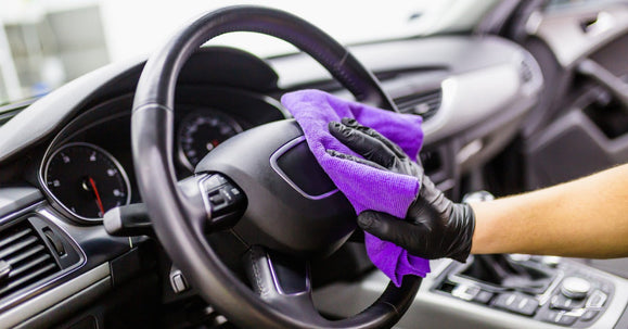 A person wearing a black glove wipes a car steering wheel with a purple cloth inside a modern vehicle.