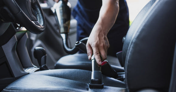 A detailer uses a handheld vacuum to clean the leather car seat interior, focusing on the center console and seat surface.