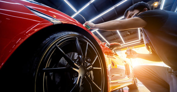 A car detailer wearing a black shirt applies a product to a red car’s front fender under bright shop lights.