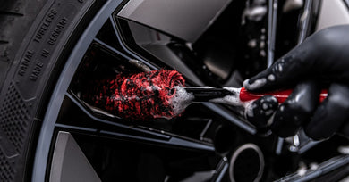 A gloved detailer scrubs a black alloy wheel with a red and black brush while soap covers the handle of the brush.